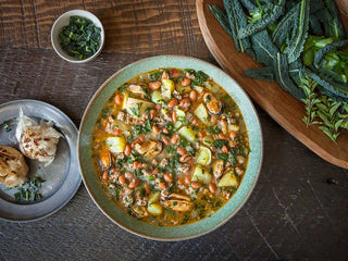 A large bowl of stew with chunks of potatoe, beans, kale, and mussels with roasted garlic on the side